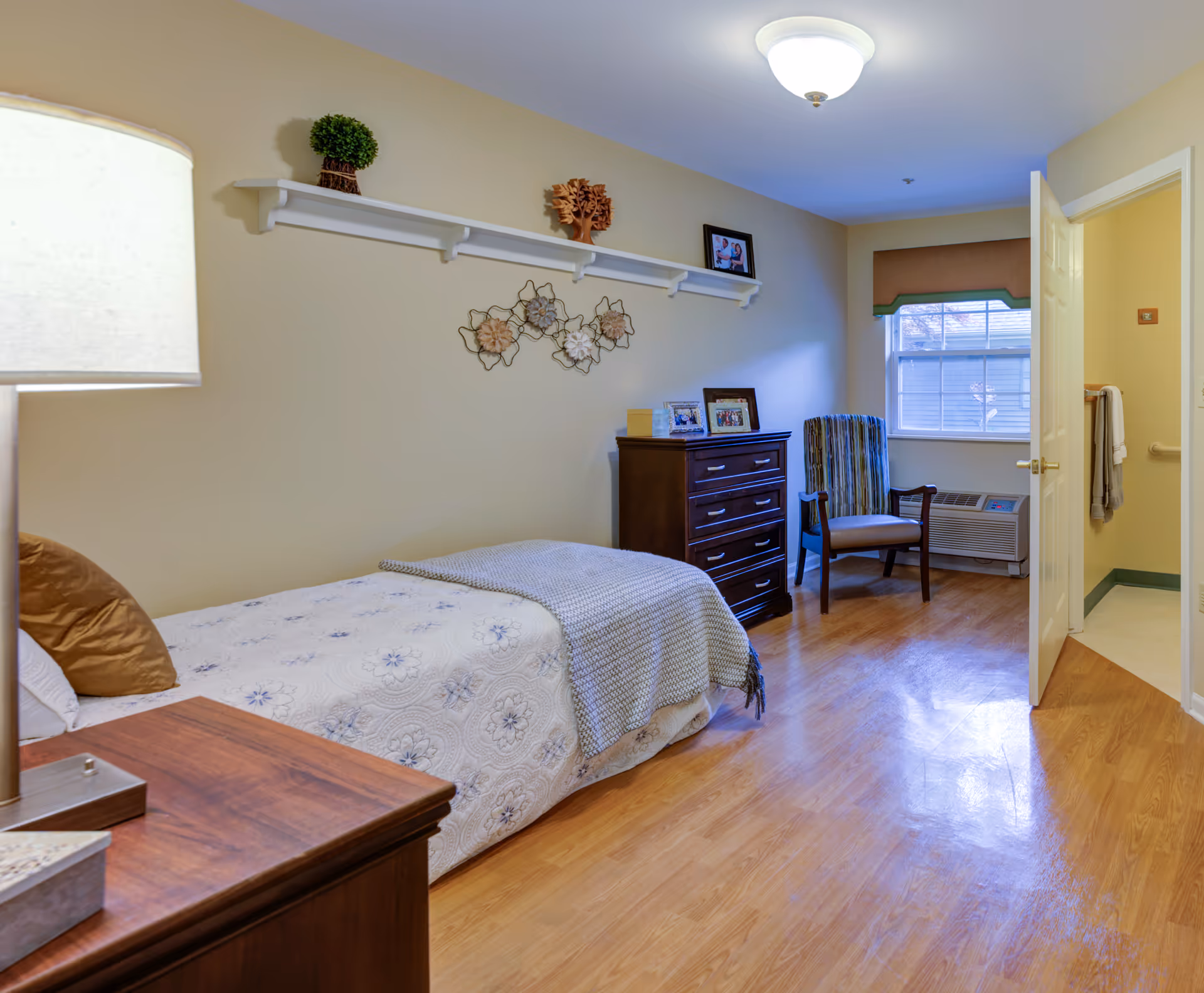 A cozy bedroom with a single bed covered in a white floral bedspread and a gray knitted throw blanket. There is a wooden nightstand with a lamp and a tissue box next to the bed. A wooden dresser with framed photos on top is against the wall, and a striped armchair is positioned near a window with a brown valance. The door to a bathroom is open, showing a towel hanging inside. The room has light-colored walls and wooden flooring.