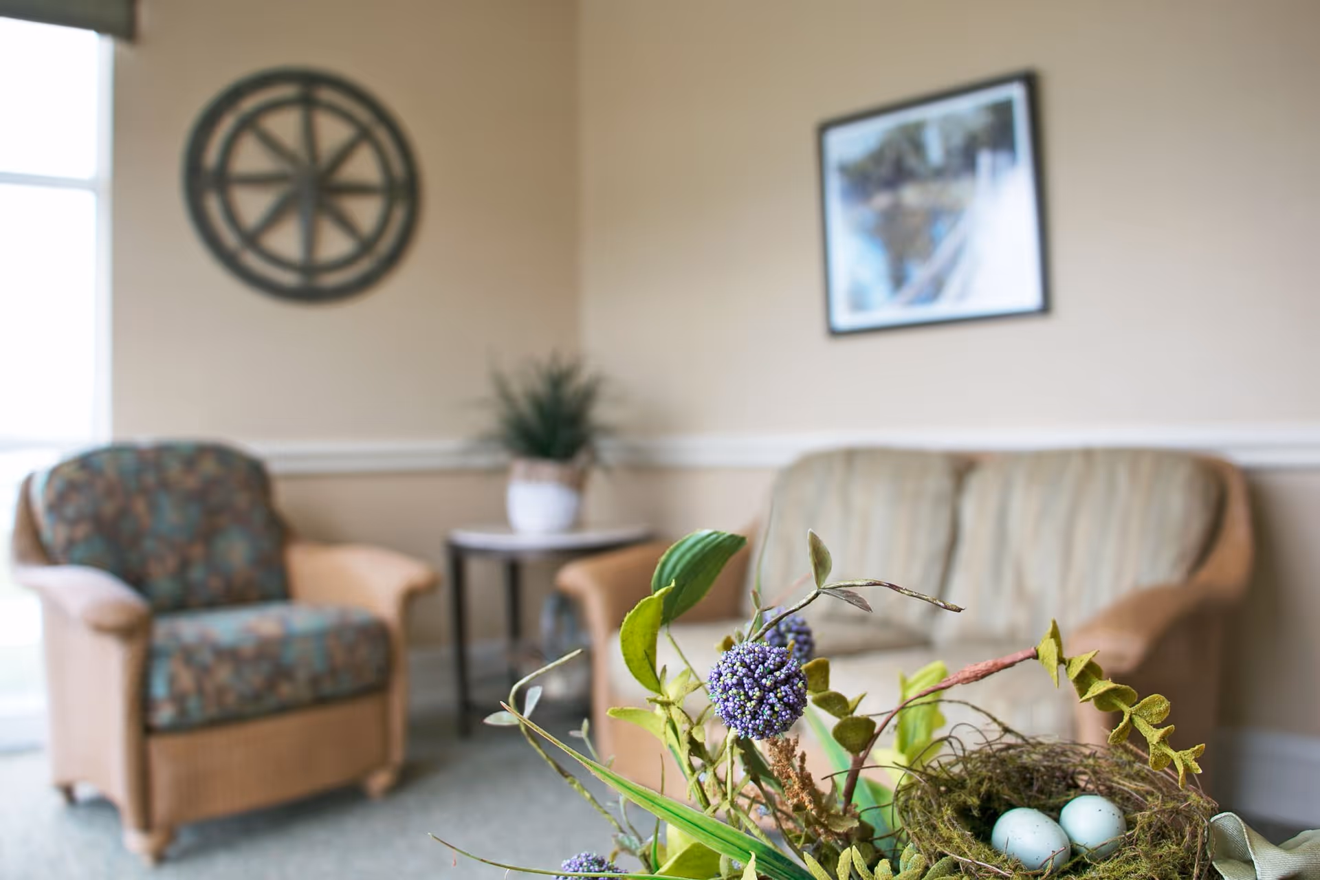 A cozy living room area with a wicker armchair with patterned cushions, a matching loveseat, a small round side table with a potted plant, and wall decorations including a circular wooden piece and a framed picture. In the foreground, there is a decorative arrangement featuring purple flowers and a small nest with two blue eggs.