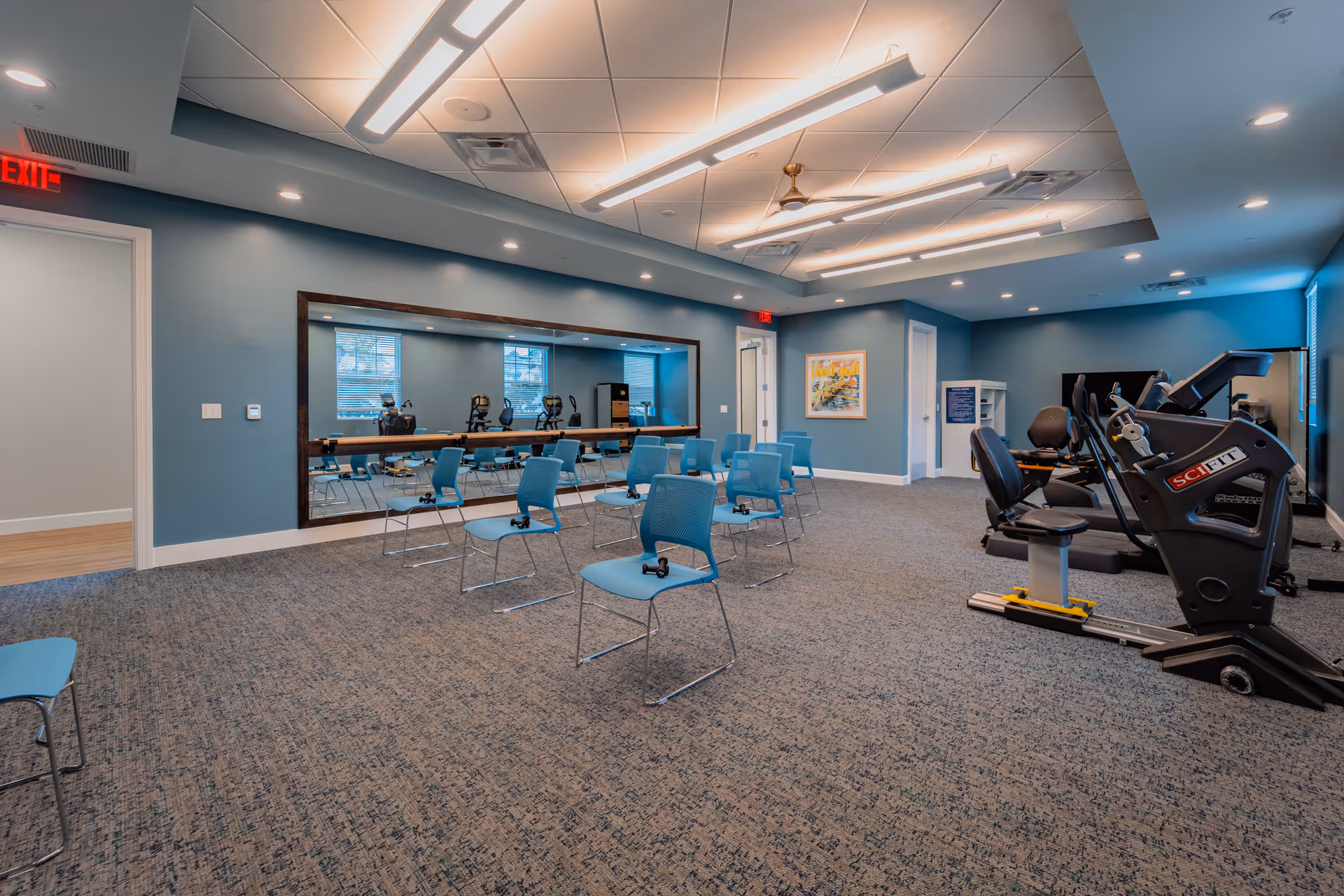 A spacious exercise room with blue walls and carpeted floor. Several blue chairs are arranged in rows facing a large wall mirror. On the right side, there are exercise machines including a recumbent bike. The room is well-lit with ceiling lights and has windows letting in natural light. There is an exit sign above a door on the left side.