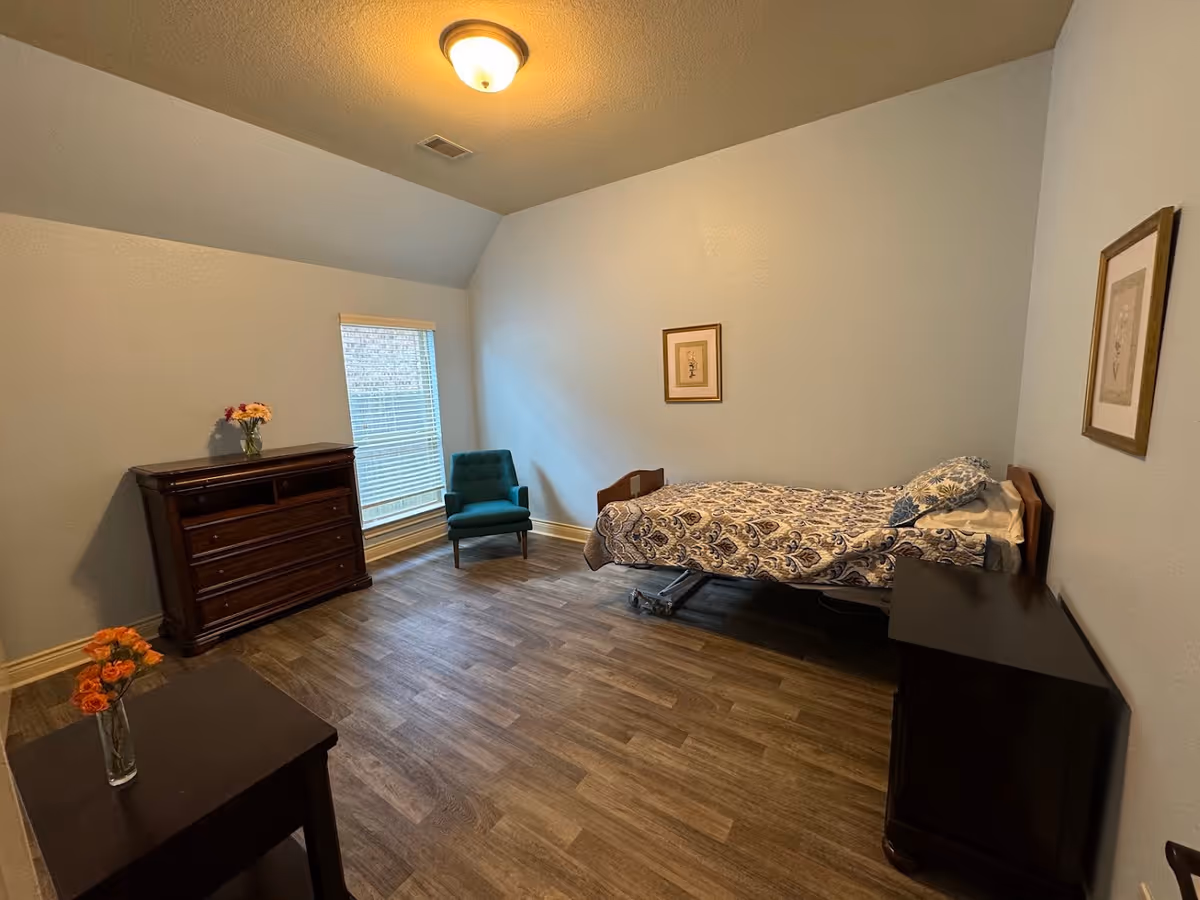 A simple bedroom with a single bed covered in a patterned bedspread, a teal armchair near a window with blinds, a wooden dresser with a vase of flowers, a small wooden side table with a vase of flowers, and two framed pictures on the walls. The room has wood flooring and light-colored walls.