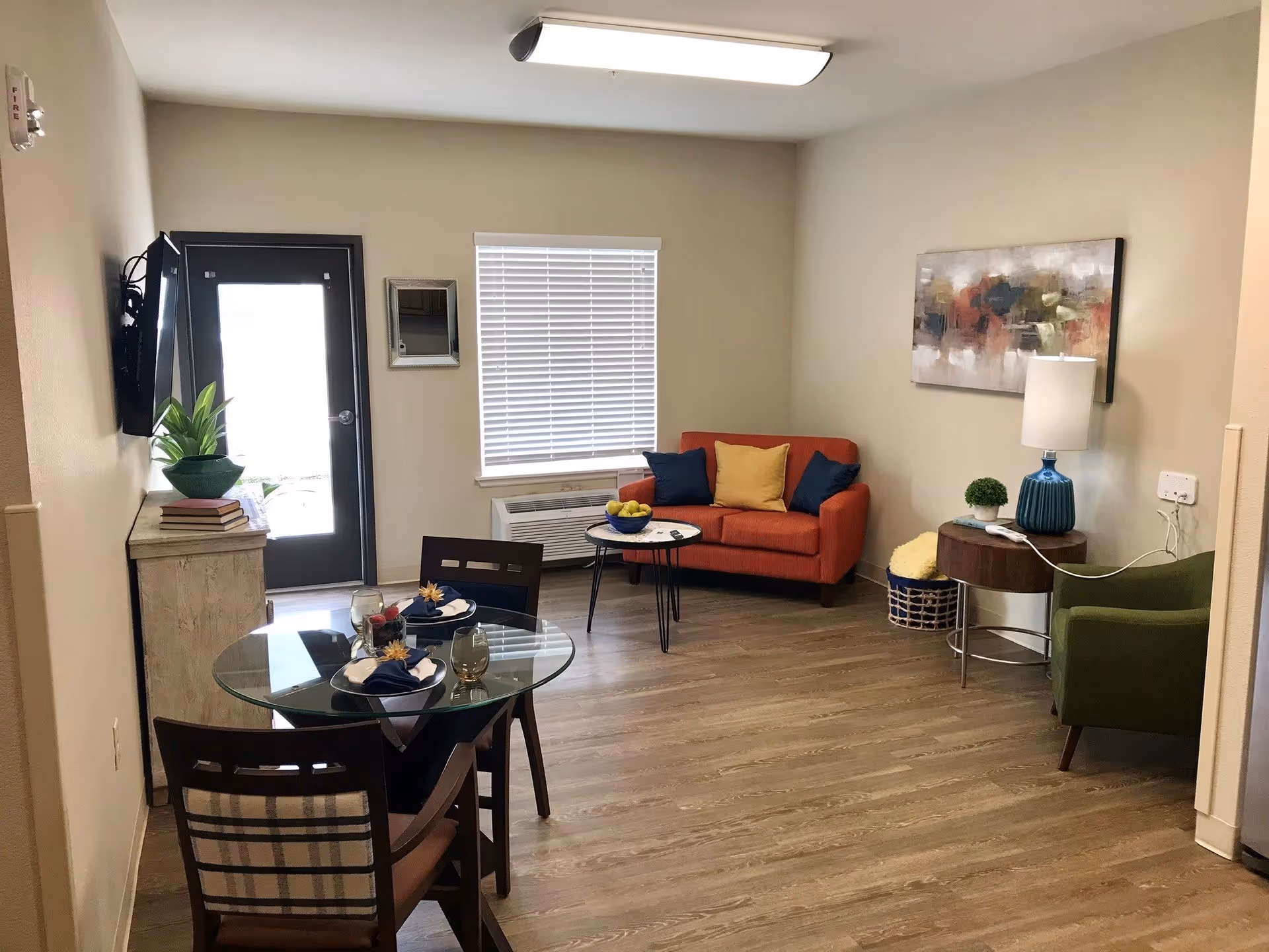 A cozy living room area with a small orange loveseat adorned with three cushions in yellow and blue. Next to it is a round wooden side table with a blue lamp and a small plant. A green armchair is positioned nearby. A round glass dining table with two chairs is set with plates, napkins, and glasses. A flat-screen TV is mounted on the wall above a wooden cabinet with a green plant and books. The room has light-colored walls, wood flooring, a window with blinds, and a door leading outside.