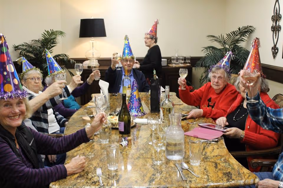 Residents wearing party hats raise glasses around a dining table.