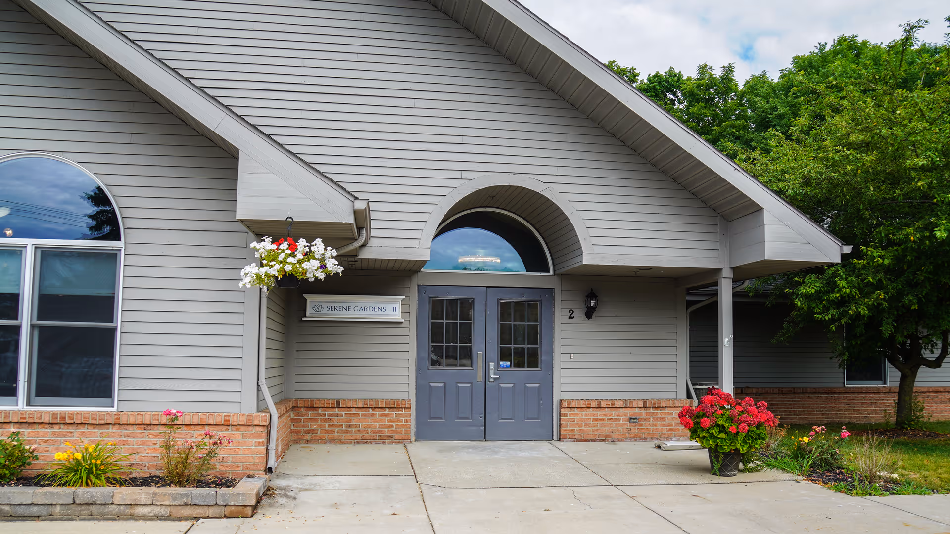 Entrance to a building with gray double doors under an arched overhang. The building has gray siding with a brick base, a hanging flower basket with white and red flowers, and a potted plant with red flowers near the entrance. There is a sign next to the door that reads 'Serene Gardens - II'.