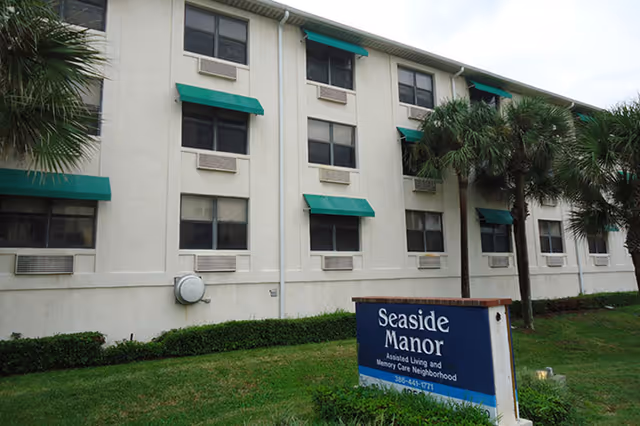 Front exterior of a three-story beige assisted living building with green window awnings, palm trees, and a 'Seaside Manor' sign on the lawn.