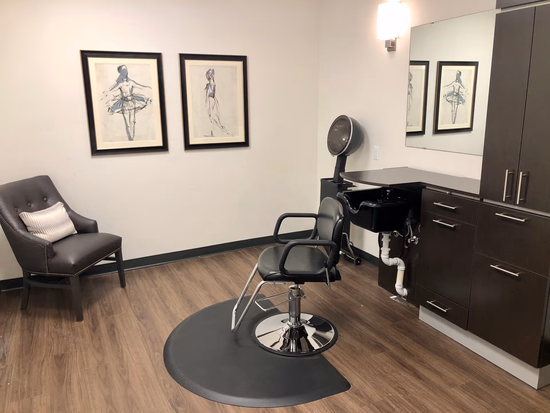 Small salon-style room with a styling chair and sink, dark cabinets, a lounge chair, and framed artwork on the wall.