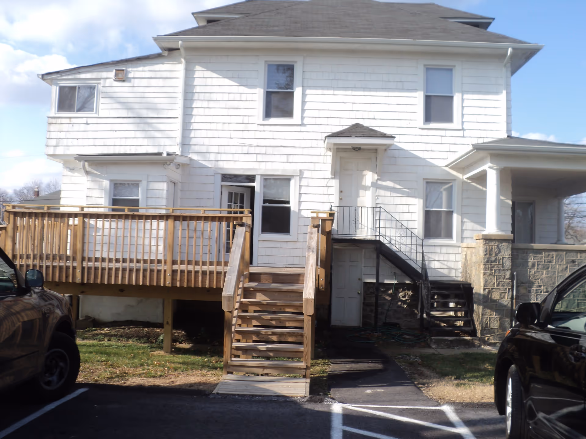 Front exterior of a white two-story house with a wooden deck, stairs, and parked cars in the driveway.
