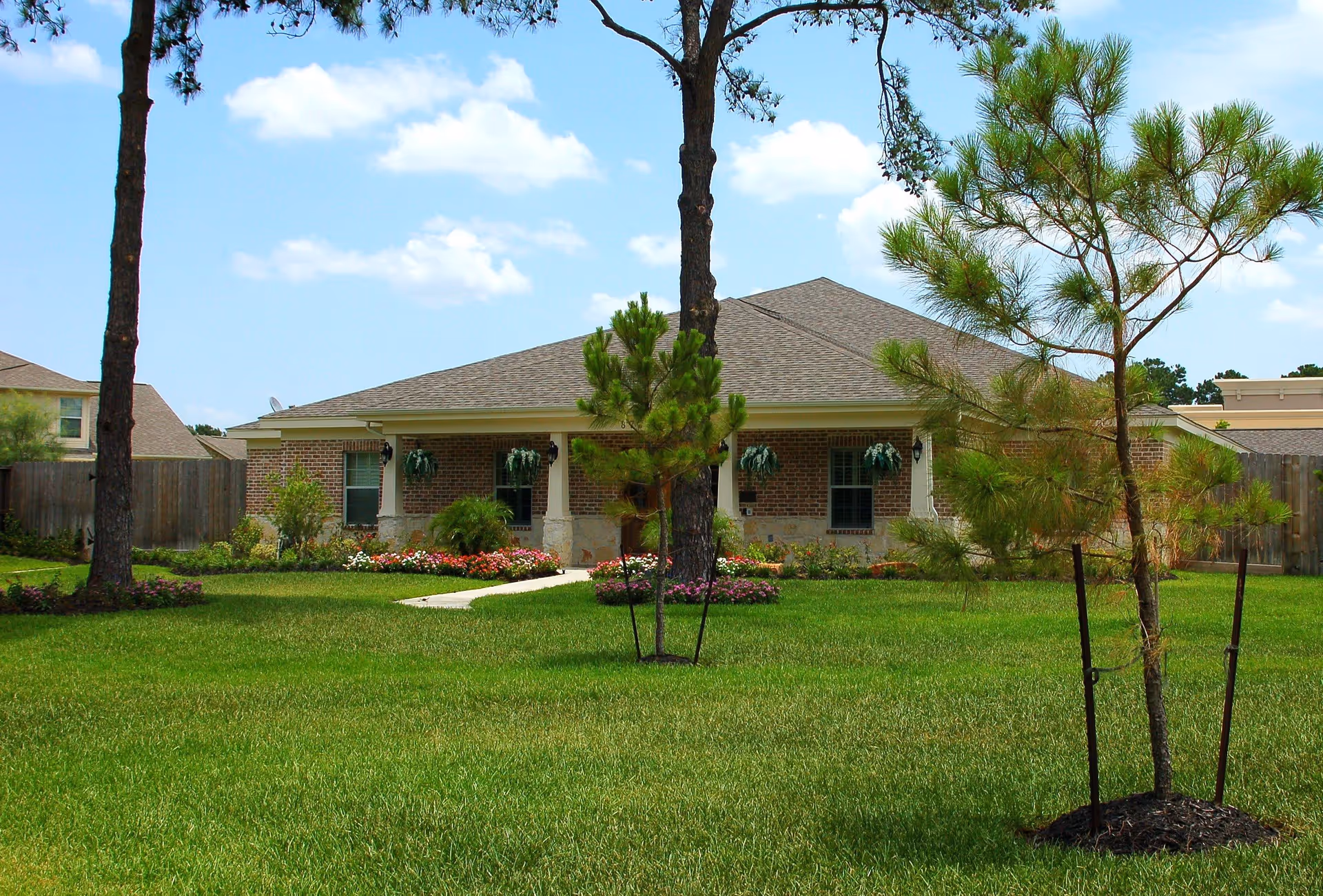 A single-story brick building with a gray shingled roof, surrounded by a well-maintained green lawn and several young trees. There are flower beds with colorful flowers near the building's entrance, and a concrete pathway leads to the front door. The sky is blue with some scattered clouds.