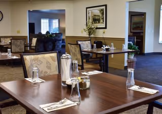 A dining area in a senior living facility with wooden tables set with napkins, silverware, glasses, and condiments. The room has beige walls with framed artwork and a carpeted floor. In the background, there is a seating area with armchairs and a window with blinds.