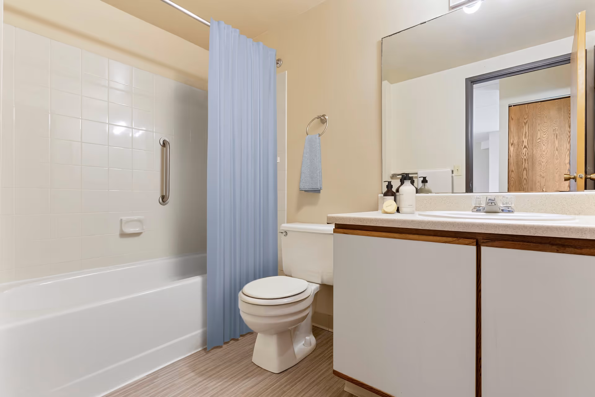 A clean bathroom featuring a white bathtub with a blue shower curtain, a white toilet, a countertop with a sink, soap dispensers, and a large mirror reflecting a wooden door and part of an adjacent room.
