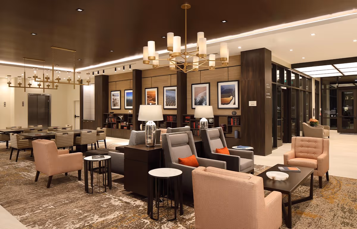 A spacious and elegantly furnished senior living facility common area with multiple seating arrangements including armchairs and tables. The room features warm lighting with modern chandeliers, framed artwork on the walls, built-in bookshelves, and large glass doors leading to another area.