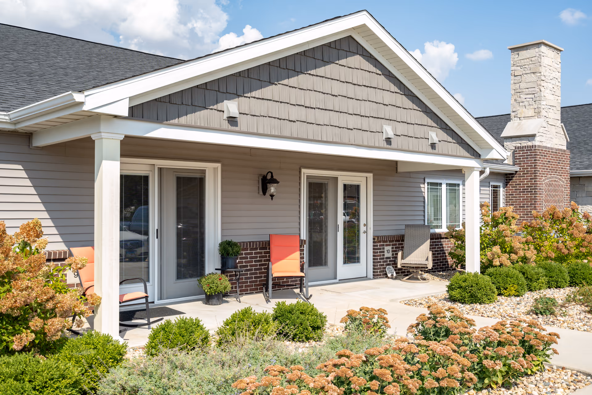 Covered patio entrance of a single-story memory care building with double glass doors, chairs, and landscaping.