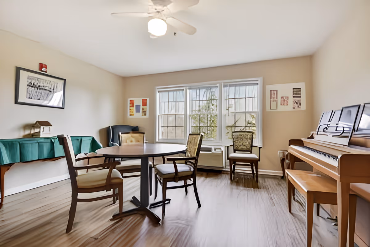 Sunlit common room with a round table and chairs, a piano, and large windows.
