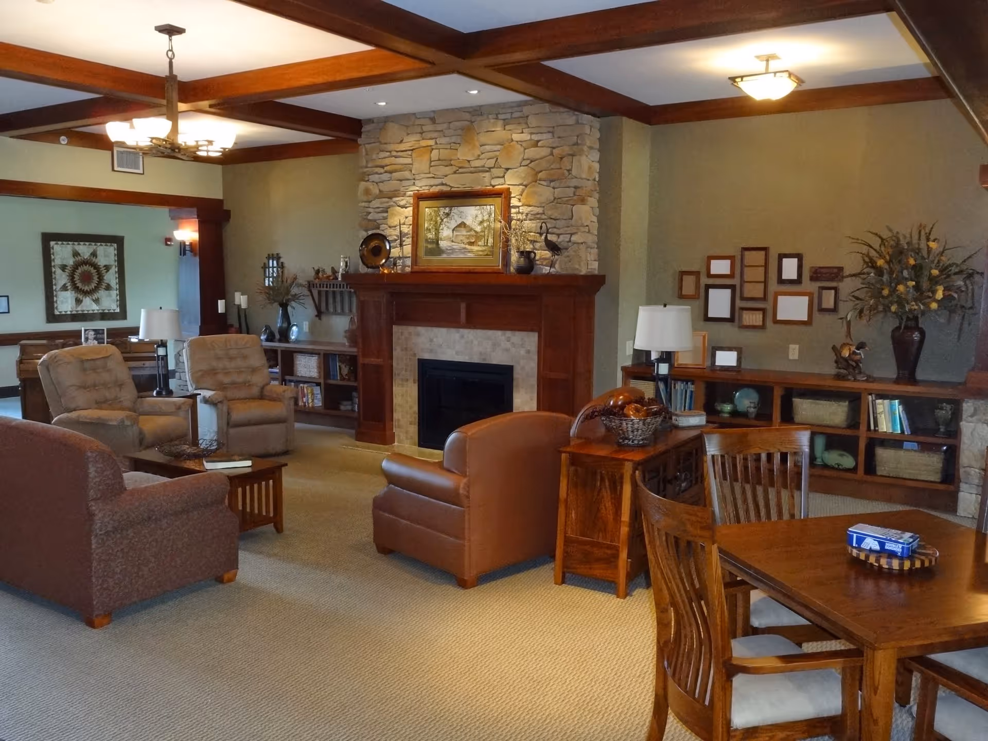 A cozy living room area in a senior living facility featuring a stone fireplace with a wooden mantel, two beige recliner chairs, a brown leather armchair, a patterned sofa, wooden coffee and side tables, and a wooden dining table with chairs. The room has a beige carpet, wooden ceiling beams, and decorative items including framed pictures, lamps, and vases with flowers.