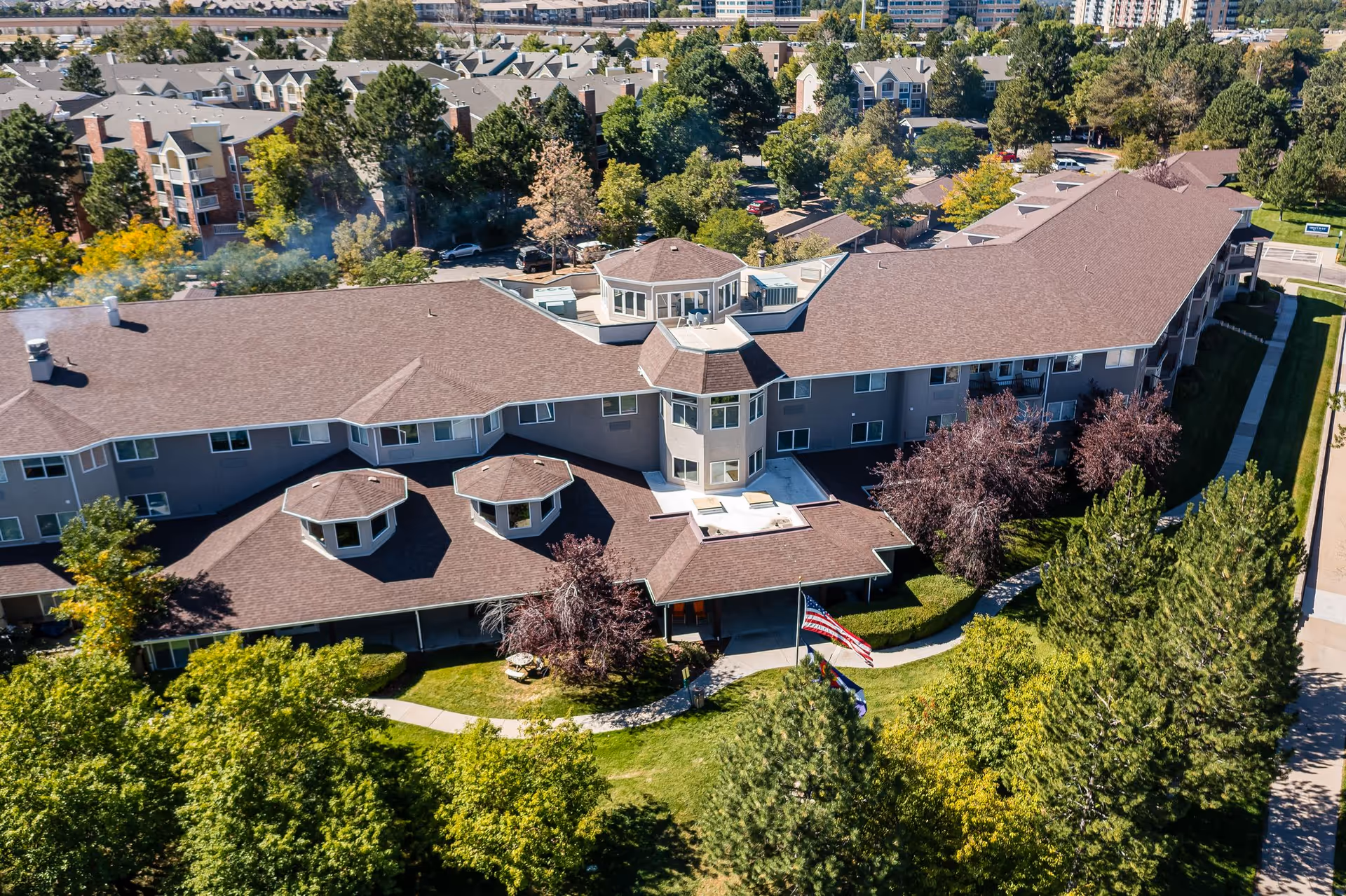 Aerial view of a large L-shaped senior living building with brown roofs, landscaped grounds, and an American flag at the entrance.