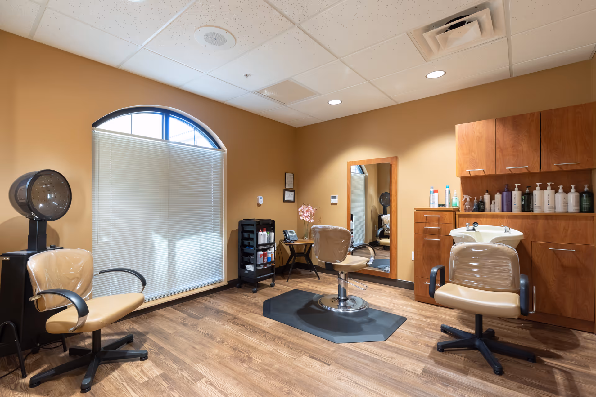 Interior salon room with styling chairs, a hair-washing sink, mirror, and cabinetry in a senior living facility.