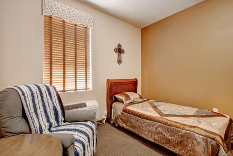 A small bedroom with a single bed covered in a patterned quilt, a cushioned armchair draped with a striped blanket, a wooden side table, a window with wooden blinds and a floral valance, and a decorative cross hanging on the wall.
