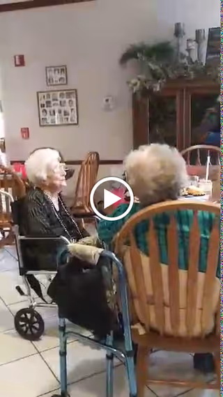 Two elderly women seated at a table in a communal dining room with a walker and a wheelchair nearby.