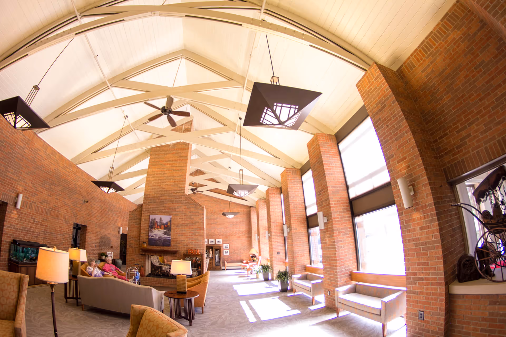 Sunlit brick-walled lounge with vaulted ceiling, couches, chairs, a fireplace and large windows in a retirement center.