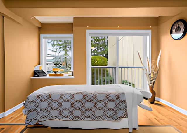 A cozy bedroom with a bed covered in a patterned blanket, a small window with a view of greenery, a sliding glass door leading to a balcony, and a decorative vase with dried branches in the corner.