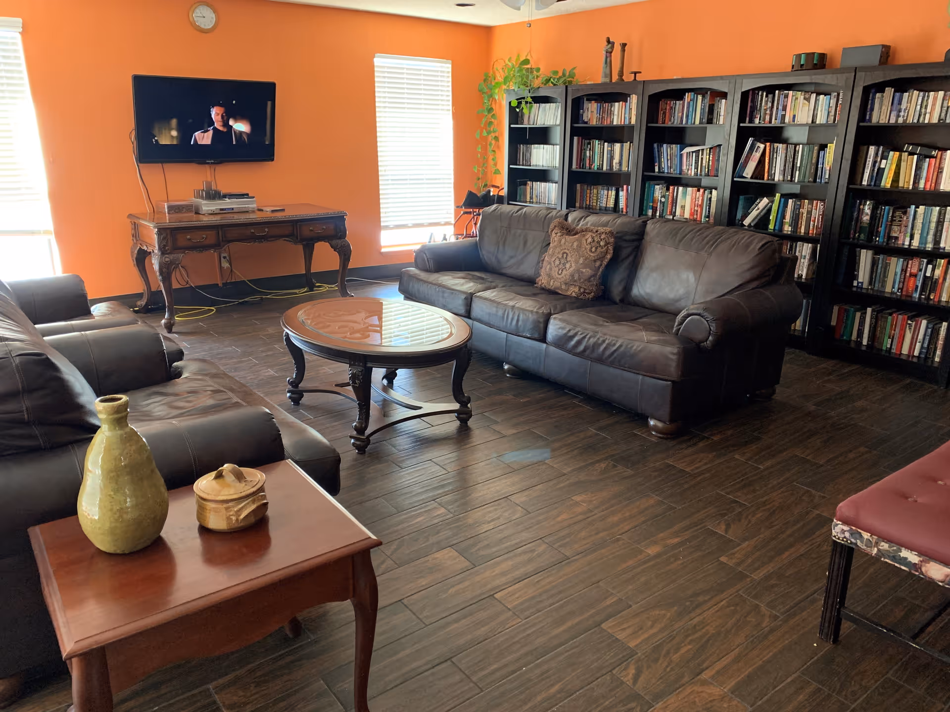 A cozy living room with orange walls, two large windows with blinds, a wall-mounted TV showing a man, two dark leather sofas with decorative pillows, a wooden coffee table with a glass top, a side table with ceramic vases, and several tall black bookshelves filled with books and some decorative items.