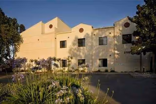 Exterior view of a beige multi-story building with several windows, surrounded by trees and flowering plants in the foreground under a clear sky.