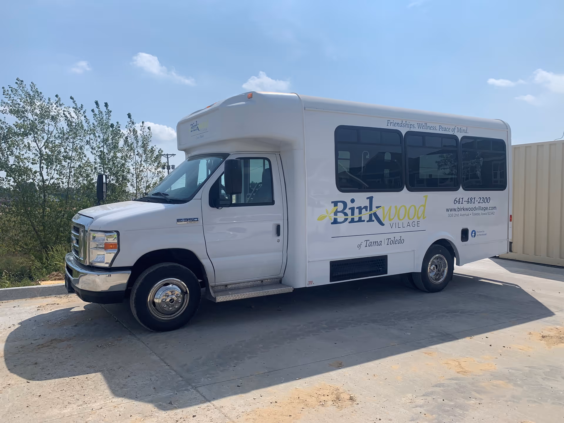 A white shuttle bus parked on a concrete surface under a blue sky with some clouds. The bus has the logo and text for Birkwood Village of Tama-Toledo on its side, along with contact information and the slogan 'Friendships. Wellness. Peace of Mind.' Trees and a beige container are visible in the background.