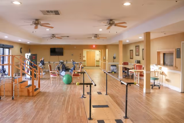 A spacious rehabilitation or physical therapy room with wooden floors, parallel bars for walking exercises, exercise equipment including stationary bikes and a green exercise ball, chairs, tables, and a small staircase with handrails. The room has ceiling fans, recessed lighting, and beige walls with framed pictures.