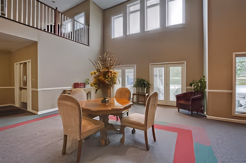 A spacious interior common area with a wooden table surrounded by four upholstered chairs. The room features high ceilings with multiple windows allowing natural light to fill the space. There are plants and floral arrangements near the windows and a wooden chair in the corner. The walls are painted beige with white trim, and the carpet has a pattern with red and green accents.