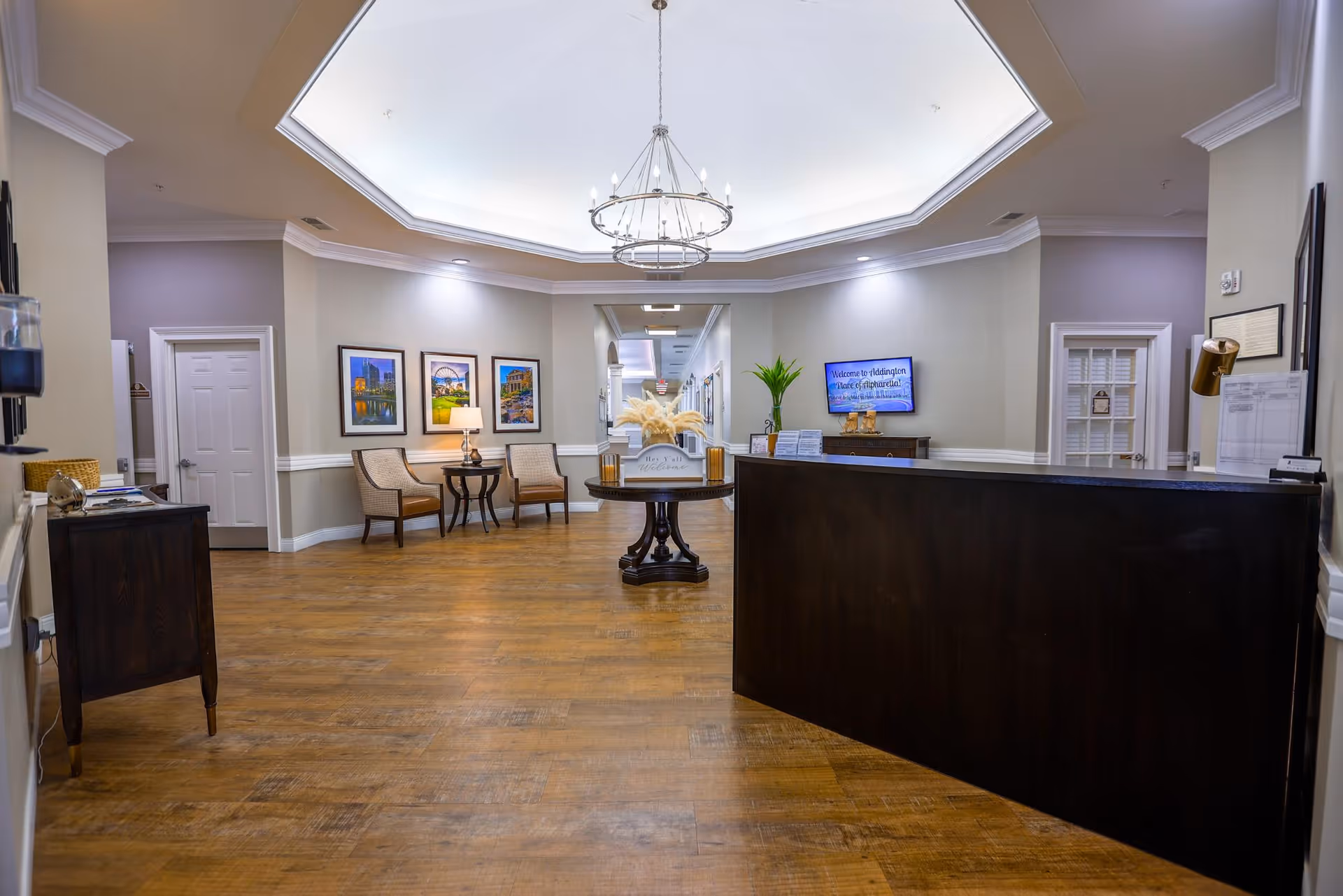 A spacious and well-lit reception area with a dark wooden front desk on the right, a round table with decorative plants in the center, and two chairs with a small table and lamp against the wall. The walls are painted light gray with white trim, and there are framed pictures hanging on the wall. A chandelier hangs from the ceiling, and a hallway extends in the background.