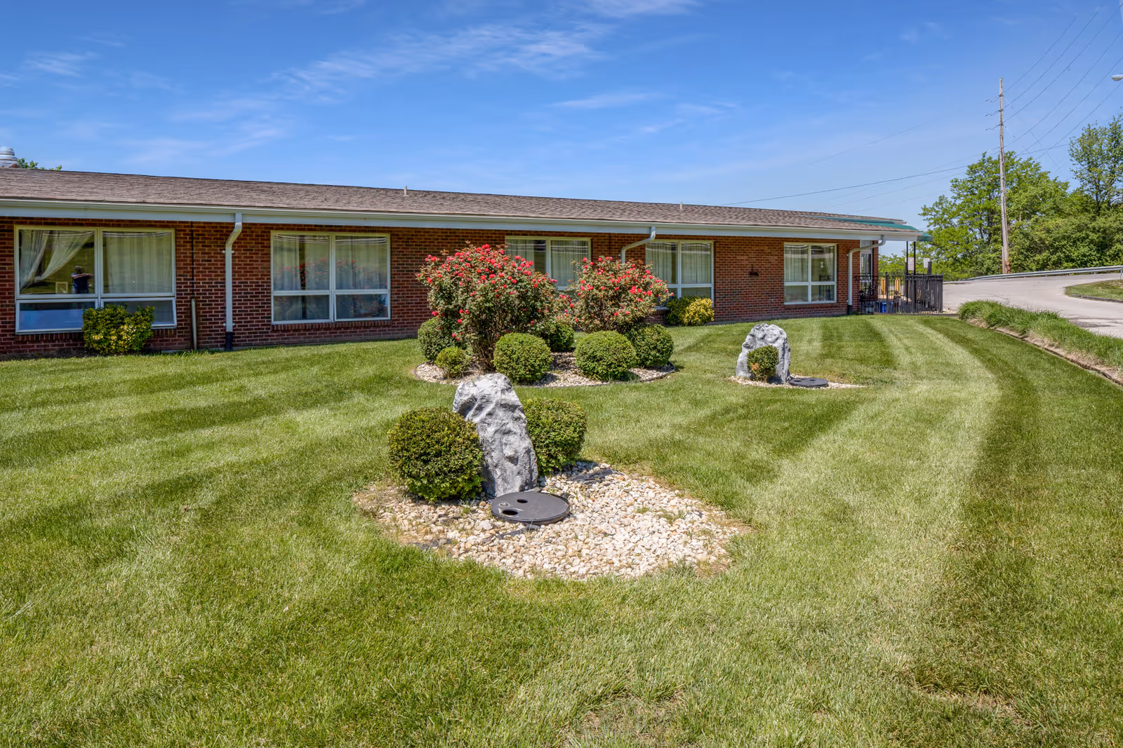A well-maintained lawn with neatly trimmed bushes and flowering shrubs in front of a single-story brick building under a clear blue sky.