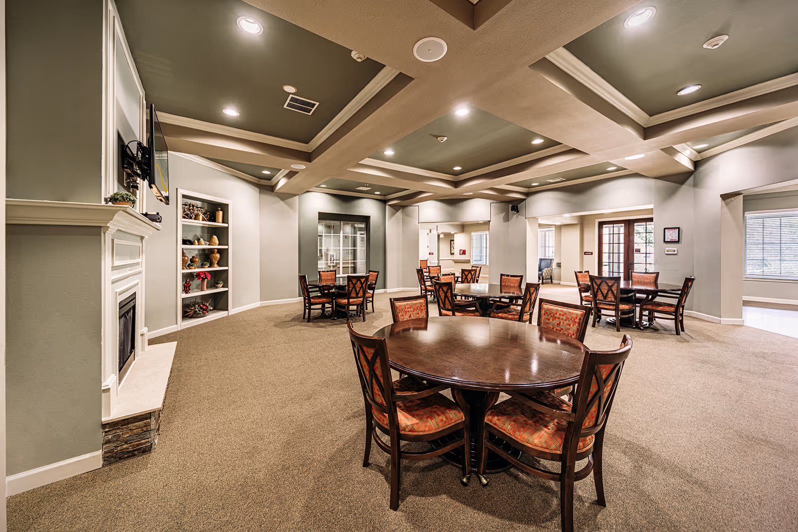 Spacious common area with multiple round wooden tables and chairs with patterned cushions, beige carpet, a white fireplace on the left wall, built-in shelves with decorative items, and a coffered ceiling with recessed lighting.