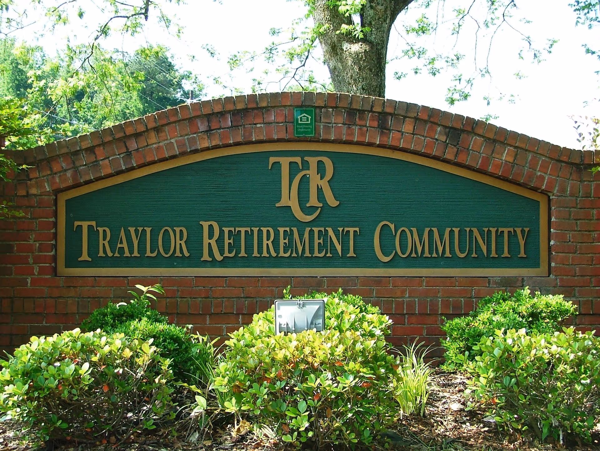 A brick sign with a green background and gold lettering that reads 'Traylor Retirement Community' with the initials 'TCR' above the text. The sign is surrounded by green bushes and trees in the background.