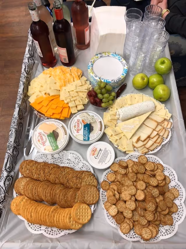 A table set with a variety of cheeses, crackers, grapes, green apples, three bottles of beverages, plastic cups, napkins, and three containers of spreadable cheese. The table is covered with a gray tablecloth and a decorative black and white runner on the side.
