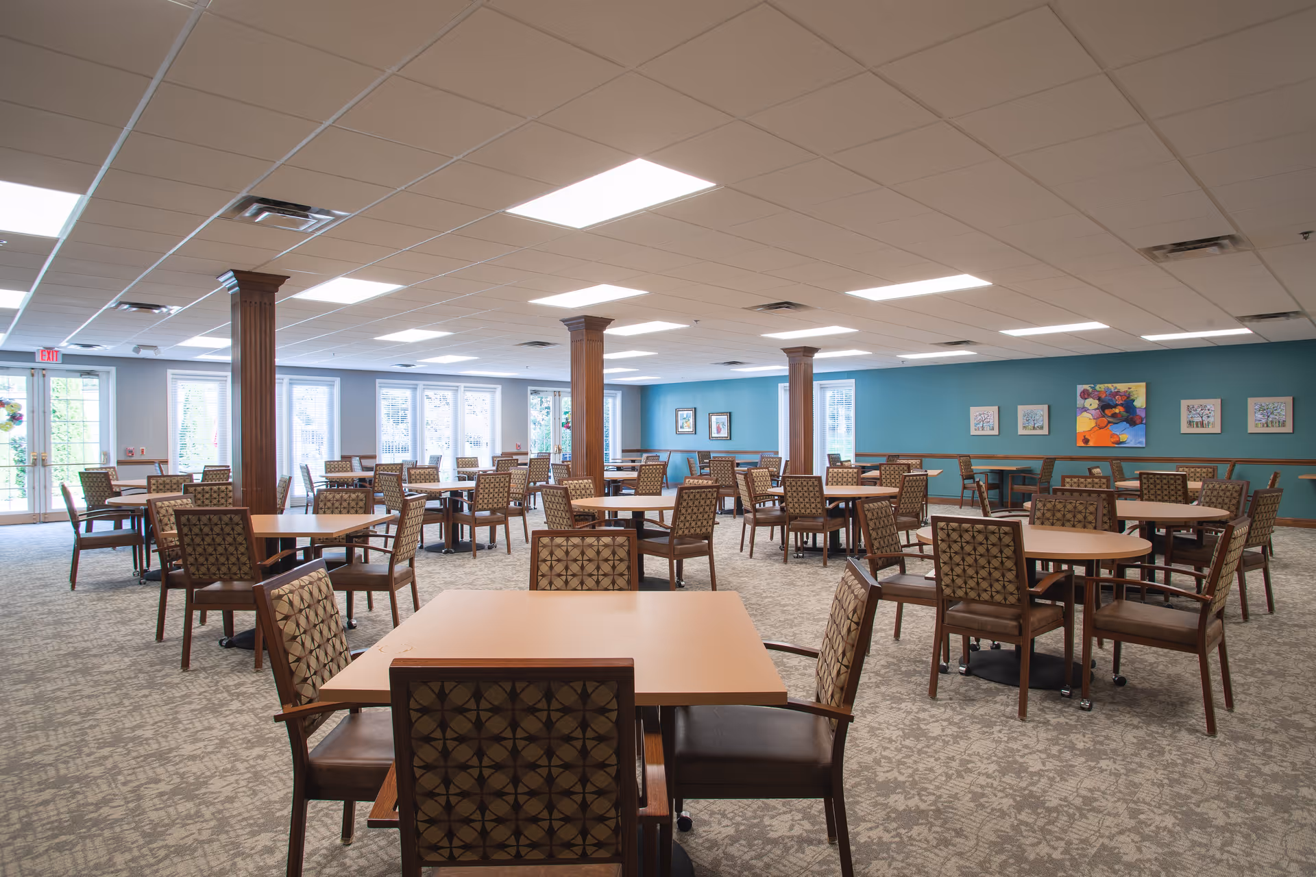 Spacious dining room with multiple round and square tables and patterned chairs, teal accent wall, and large windows letting in natural light.