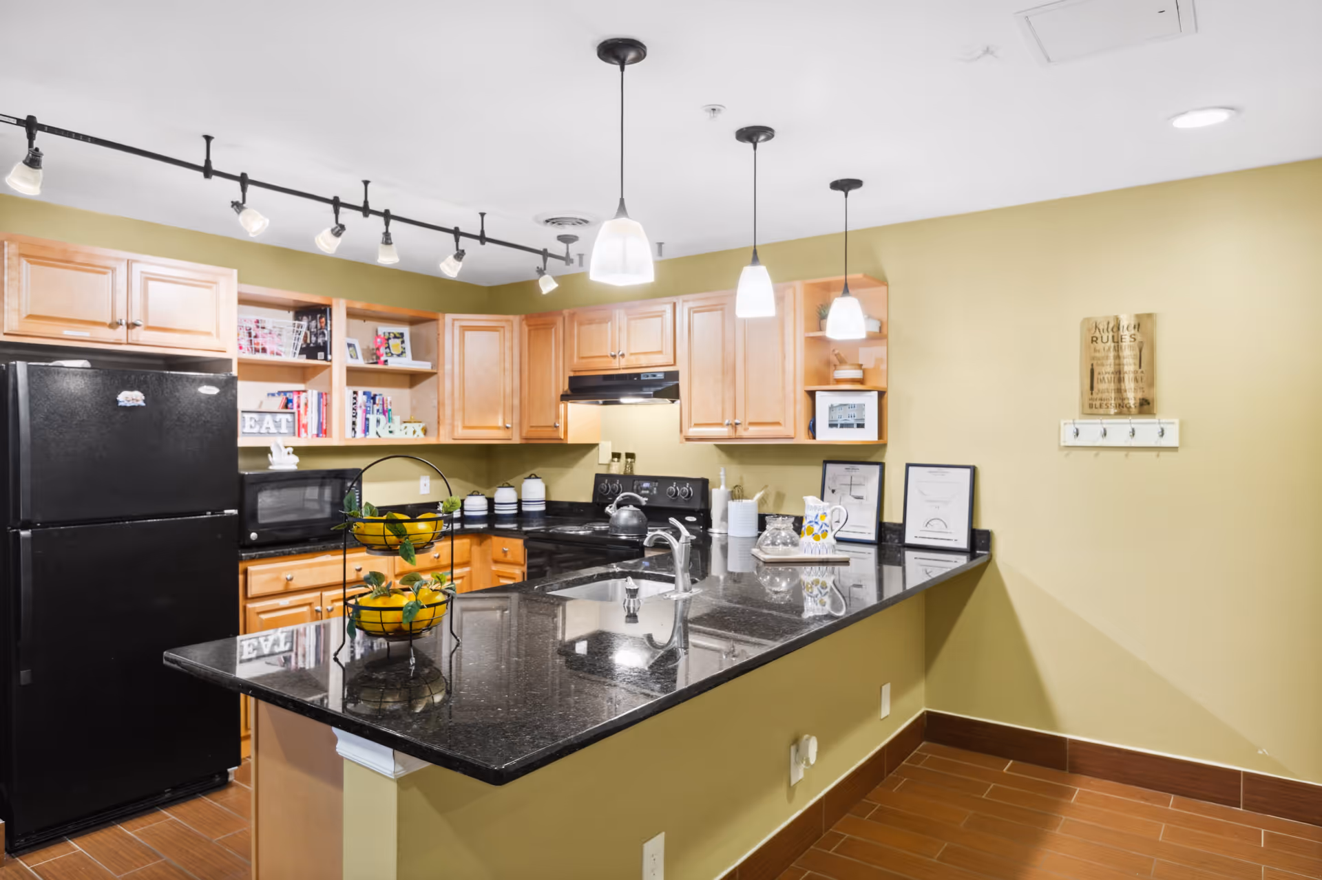 A modern kitchen with light wood cabinets, black granite countertops, and a black refrigerator. The kitchen features a double sink, a black stove with a kettle, a microwave, and open shelves with decorative items and books. Three pendant lights hang above the countertop, and the walls are painted a light yellow-green color.