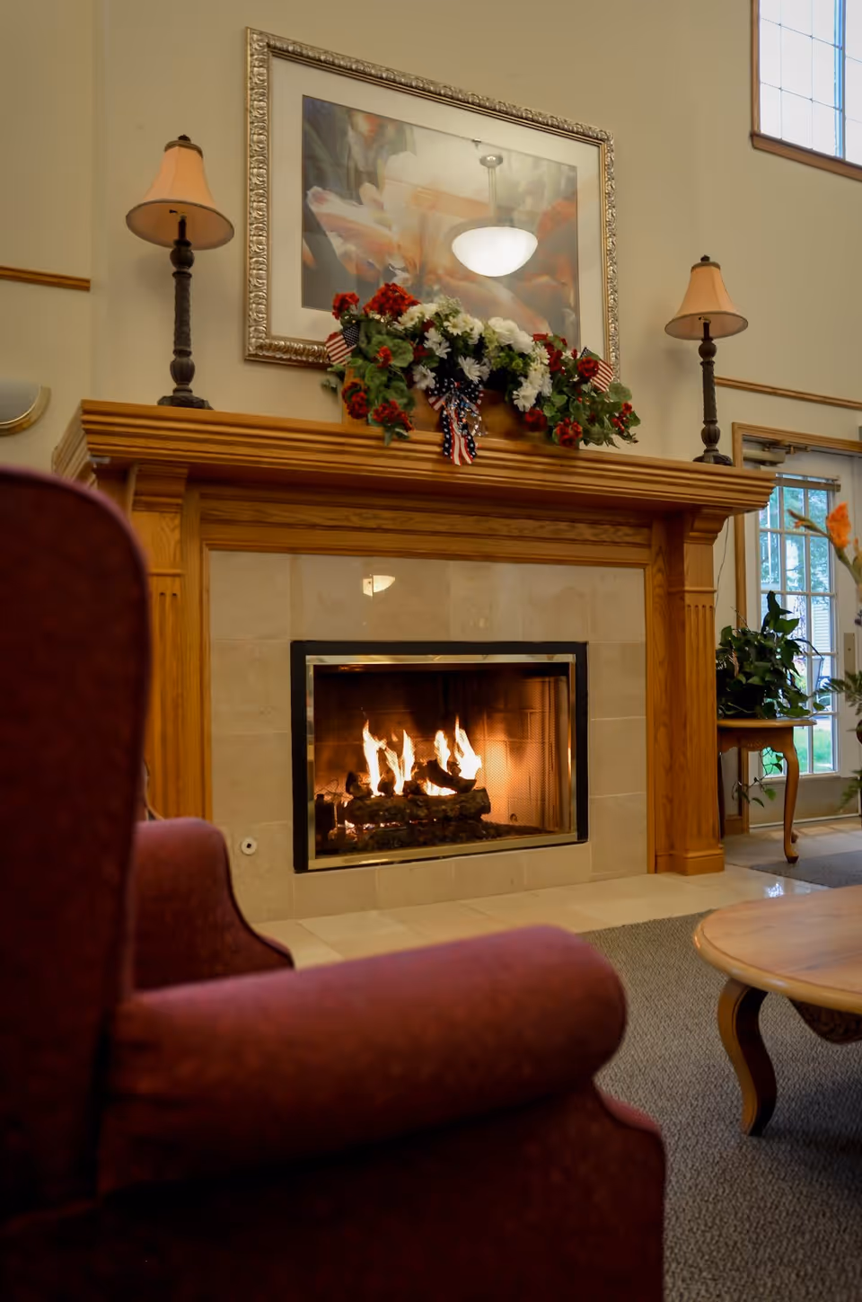 A cozy living room area featuring a lit fireplace with a wooden mantel decorated with a floral arrangement and small American flags. Above the mantel is a framed painting. Two table lamps with beige shades are placed on either side of the mantel. A red upholstered armchair is partially visible in the foreground, and a wooden coffee table and potted plants are seen in the background near large windows.