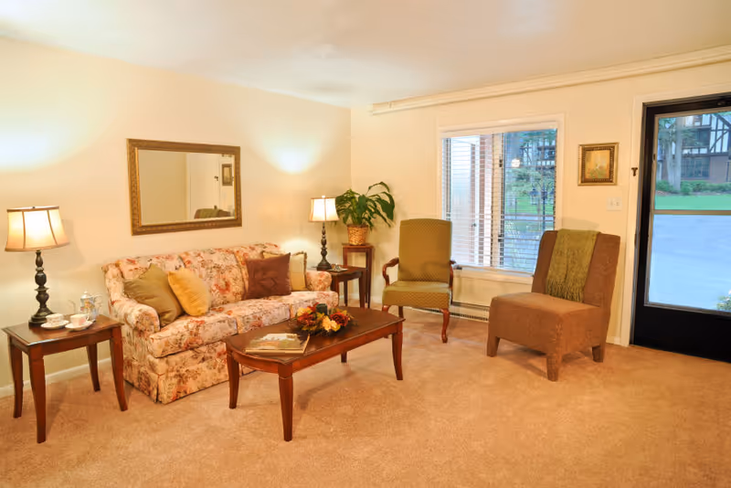 A cozy living room with a floral patterned sofa adorned with multiple cushions, two armchairs, a wooden coffee table with a decorative centerpiece and a book, two side tables each with a lamp, a large mirror on the wall, a potted plant on a small stand, a window with blinds, and a glass door leading outside.