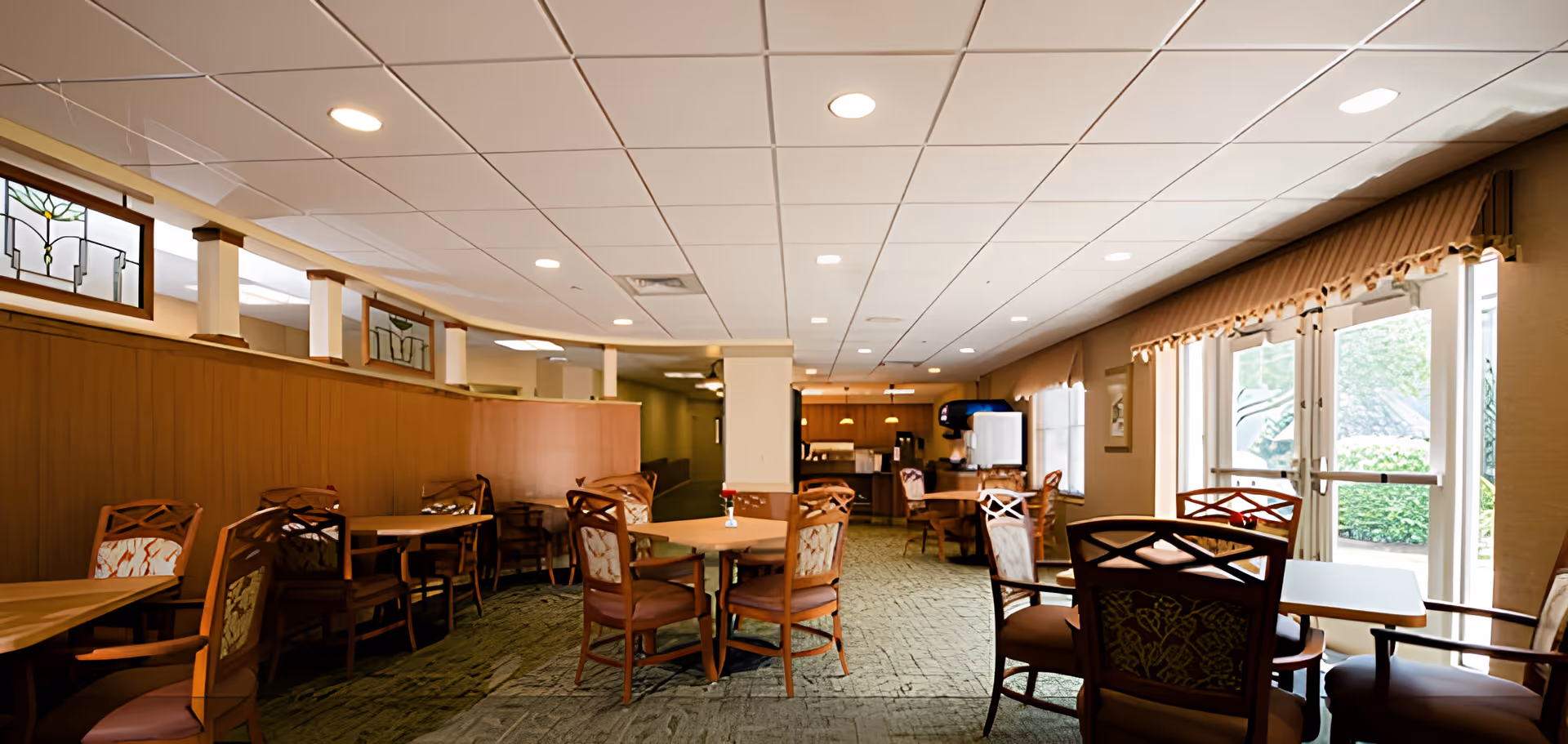 A well-lit dining area in a senior living facility with multiple wooden tables and chairs arranged neatly. The room features large windows with beige curtains allowing natural light to enter, and decorative stained glass panels above a wooden partition. The far end of the room has a beverage station with a soda fountain and other amenities.