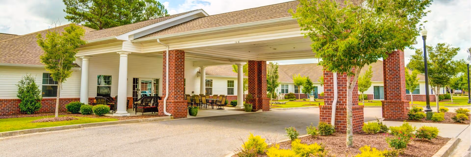 Covered porte-cochere entrance of a single-story senior living facility with brick columns, outdoor seating, and landscaped grounds.