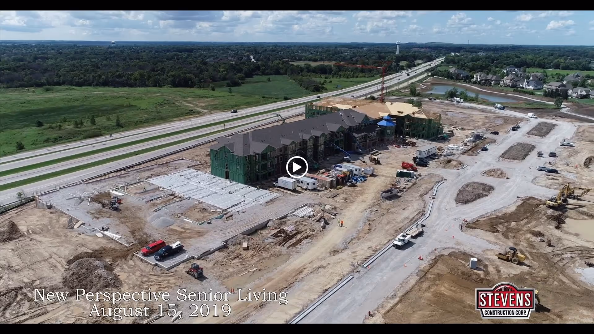 Aerial view of a senior living building under construction next to a highway with construction equipment and cleared lots.