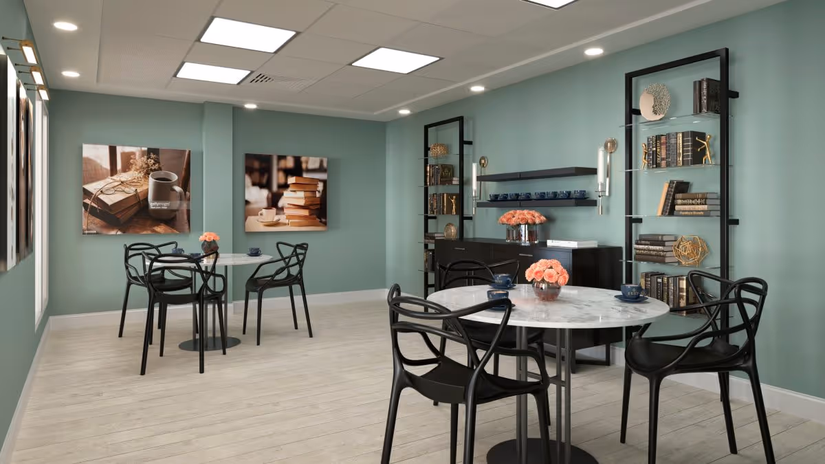 A cozy dining area with two round marble tables, each surrounded by four black chairs. The walls are painted a soft green and decorated with framed pictures of books and coffee cups. There are two black shelving units filled with books and decorative items, and a black sideboard with shelves holding blue cups and a vase of pink flowers. The floor is light wood, and the ceiling has recessed lighting and square light panels.