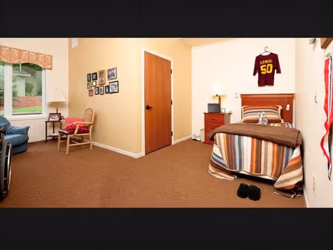 A cozy bedroom with a single bed covered in a striped blanket and a brown throw. A maroon sports jersey with the name 'Lerner' and number 50 hangs on the wall above the bed. Next to the bed is a wooden nightstand with a lamp and a small radio. A pair of black slippers is on the carpeted floor near the bed. On the left side of the room, there is a wooden chair with a red cushion and a small side table with a lamp. Several framed photos are hung on the beige wall near a closed wooden door. A window with a valance lets in natural light.