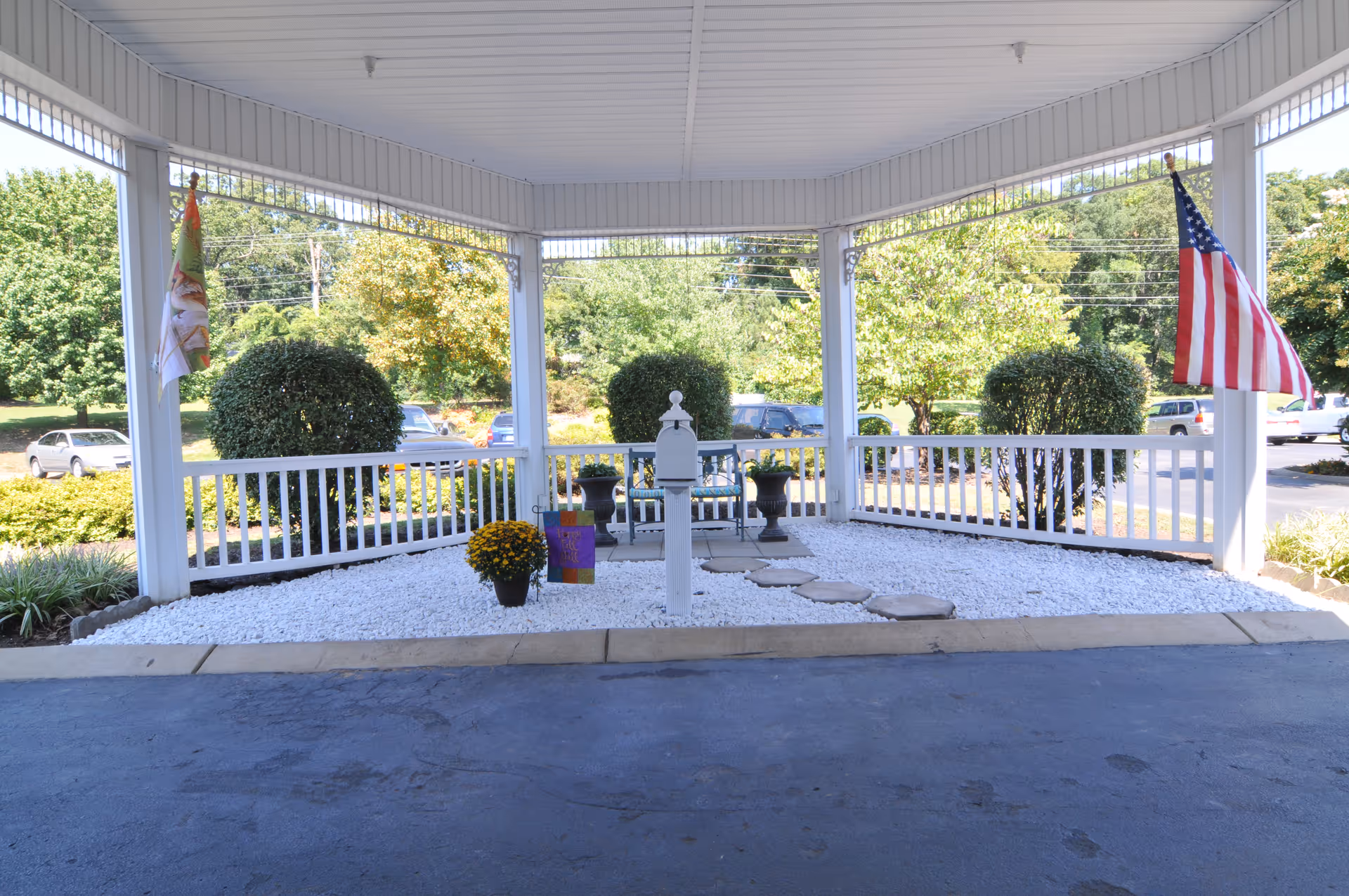Covered entrance/portico with white railing, white decorative rock landscaping, stepping stones, seating and an American flag at the front of the facility.