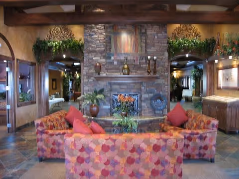 A cozy living room area with a stone fireplace as the focal point. The fireplace is decorated with candles, vases, and a piece of artwork above it. In front of the fireplace are two patterned sofas with red and orange cushions arranged around a glass coffee table with a floral centerpiece. The room features warm lighting, wooden beams on the ceiling, and greenery accents above the fireplace and in the background.