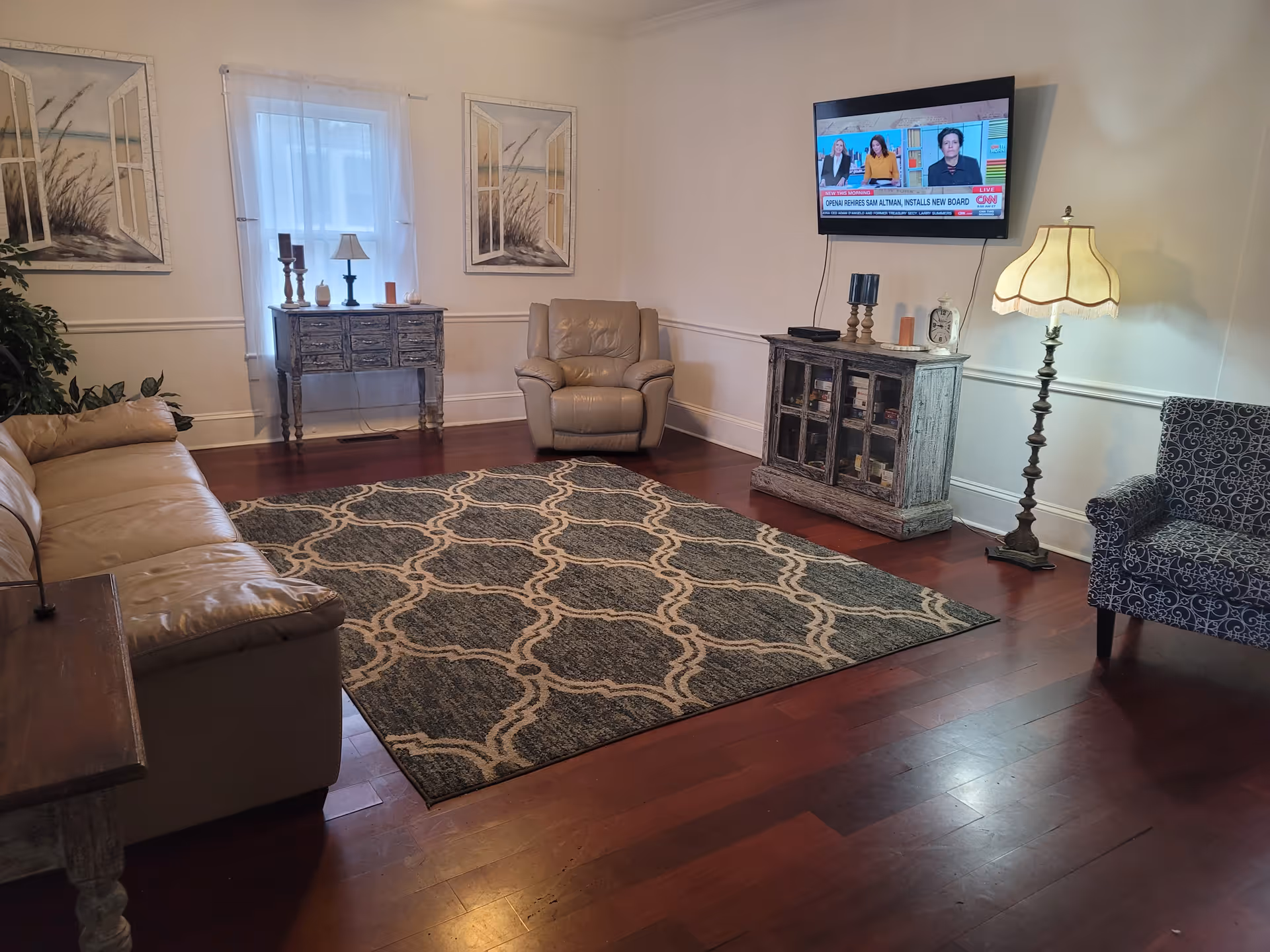 A cozy living room with a beige leather sofa on the left, a patterned armchair on the right, and a beige recliner chair in the center. A large patterned rug covers the wooden floor. On the far wall, there is a window with sheer white curtains flanked by two framed pictures. Below the window is a small rustic wooden table with decorative items. A flat-screen TV is mounted on the right wall above a distressed wooden cabinet with glass doors. A tall floor lamp with a cream-colored shade stands next to the cabinet.