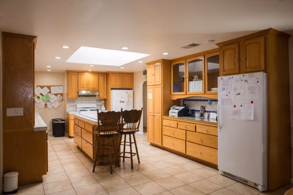 A spacious kitchen with wooden cabinets and drawers, a white refrigerator covered with papers, a white stove, and a kitchen island with two wooden chairs. The floor is tiled, and there is a skylight in the ceiling providing natural light. Various kitchen appliances and items are visible on the countertops.