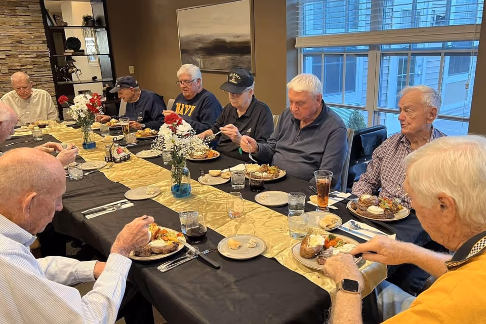 A group of elderly men sitting around a long dining table enjoying a meal together in a well-lit room with large windows. The table is covered with a black tablecloth and a gold table runner, decorated with vases of flowers. Plates of food and drinks are placed in front of each person.