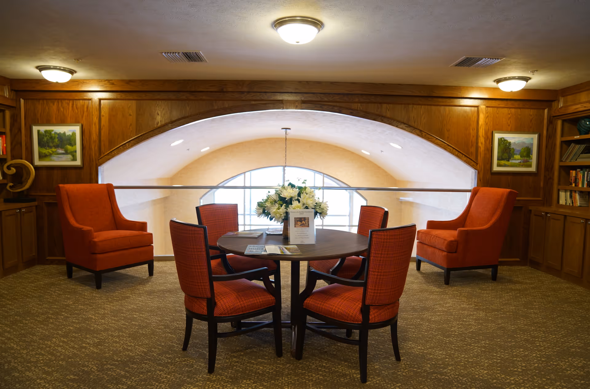 A cozy seating area in a retirement community with a round wooden table surrounded by four red cushioned chairs. Two matching red armchairs are placed against wood-paneled walls with built-in bookshelves and framed landscape paintings. A floral centerpiece is on the table, and a large arched window with a railing is in the background.