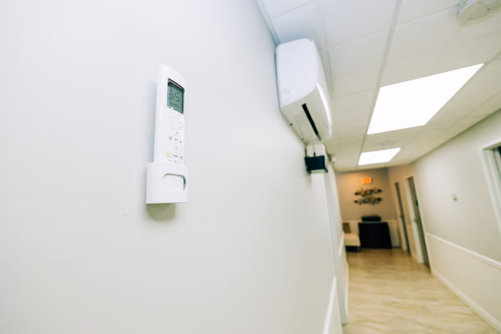 A clean, well-lit hallway with light-colored walls and wood flooring. A white air conditioning unit and its remote control are mounted on the wall. The hallway has several doorways and ceiling lights, with a piece of wall art and furniture visible at the far end.