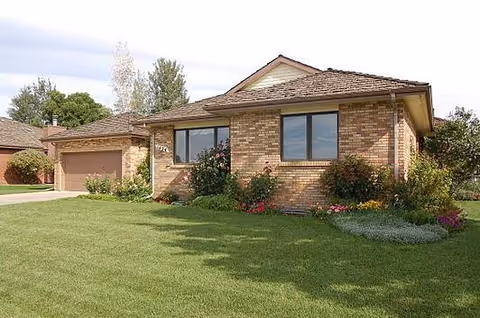 Single-story brick house with a brown shingled roof, two large windows, and a garage on the left side. The front yard has a well-maintained green lawn and various shrubs and flowers along the house's foundation.
